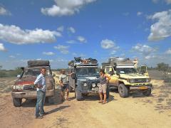 A group of travelers stands in front of three 4x4 vehicles, outfitted for overlanding, in a sunny, dusty landscape with a blue sky and puffy white clouds.